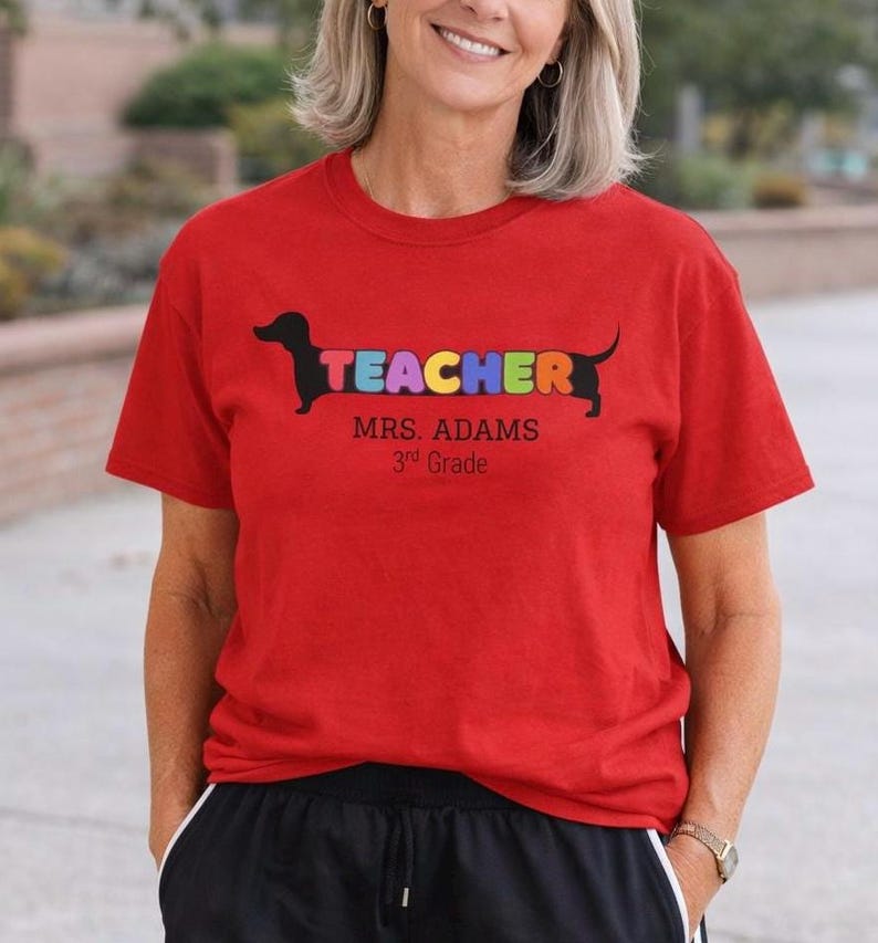 Pode incluir: Camiseta vermelha com a palavra &laquo;TEACHER&raquo; em letras coloridas, desenhada como um c&atilde;o dachshund. Abaixo, o texto &laquo;MRS. ADAMS 3rd Grade&raquo; em preto. A camiseta &eacute; de gola redonda.