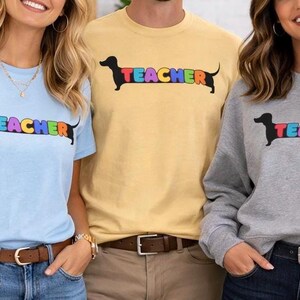 Pode incluir: Tr&ecirc;s pessoas vestindo camisas e um moletom com um desenho de cachorro salsicha. A palavra "TEACHER" &eacute; impressa em cores do arco-&iacute;ris no corpo do cachorro. As camisas s&atilde;o azul claro e amarelo, e o moletom &eacute; cinza.