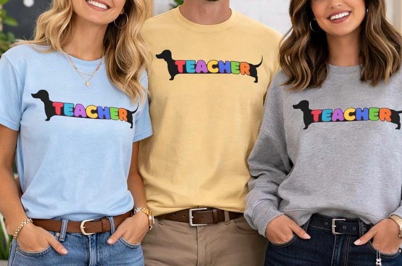 Pode incluir: Tr&ecirc;s pessoas vestindo camisas e um moletom com um desenho de cachorro salsicha. A palavra "TEACHER" &eacute; impressa em cores do arco-&iacute;ris no corpo do cachorro. As camisas s&atilde;o azul claro e amarelo, e o moletom &eacute; cinza.
