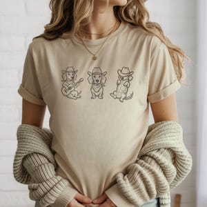 May include: A beige t-shirt featuring three dachshunds wearing cowboy hats. One dog plays a guitar, another wears a bandana, and the third reclines. The graphic is in black ink.