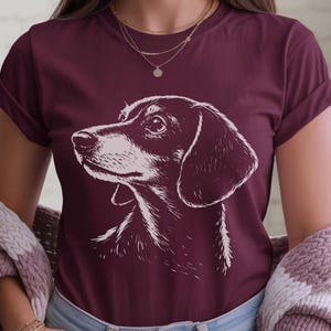 May include: Burgundy t-shirt featuring a detailed white illustration of a dachshund dog's head. The dog's profile is prominently displayed, with a focus on its face and ears. The shirt is a solid color, and the dog illustration is the main design element.