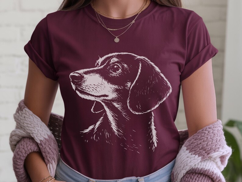 May include: Burgundy t-shirt featuring a detailed white illustration of a dachshund dog's head. The dog's profile is prominently displayed, with a focus on its face and ears. The shirt is a solid color, and the dog illustration is the main design element.