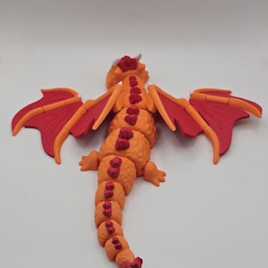 May include: A detailed orange and red dragon figurine with outstretched wings. The dragon has a segmented body, small horns, and a long tail. The wings are red with orange accents. The figurine is set against a plain white background.
