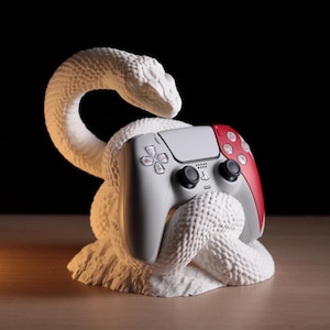 Puede incluir: Una escultura de serpiente blanca y texturizada sostiene un mando de videojuegos gris y rojo. El mando tiene botones negros y un logotipo de PlayStation. La escultura está sobre una superficie de madera sobre un fondo negro.