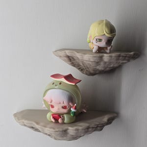 May include: Two small chibi-style figurines displayed on separate floating rock-textured shelves. The top figurine has blonde hair and green eyes. The bottom figurine wears a green hood with a red book on top and holds a small red device.