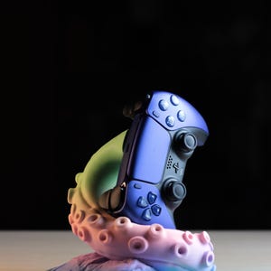May include: A blue and black video game controller rests on a colorful, tentacle-shaped stand. The stand features a gradient of colors, including pink, green, and blue. The controller has a sleek design with buttons and joysticks.
