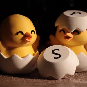 May include: A pair of yellow chick-shaped salt and pepper shakers. Each chick is emerging from a white eggshell. One eggshell has the letter "P" and the other has the letter "S". The shakers are on a wooden surface.