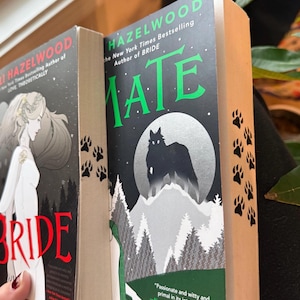 Może przedstawiać: Wyświetlane są dwie książki Hazelwood. Książka po lewej stronie nosi tytuł "Bride" z kobietą w białej sukni na okładce. Książka po prawej stronie nosi tytuł "Mate" z sylwetką wilka i sceną górską. Odbitki łap znajdują się z boku książki.