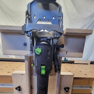 Op de afbeelding: Een zwart en grijs elektrisch gereedschap met groene accenten, gemonteerd op een houten frame. Het gereedschap heeft een cilindrisch lichaam en een grote, schuine kop. Het woord "DOMINO" is zichtbaar aan de zijkant. Het gereedschap is ontworpen voor houtbewerking.