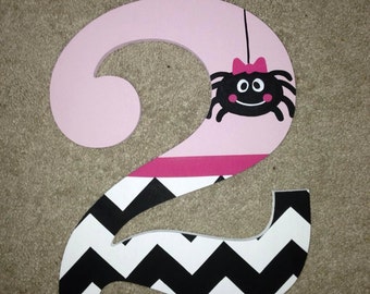 May include: Pink wooden number two with a black and white chevron pattern. A black spider with a pink bow is on the top of the number.