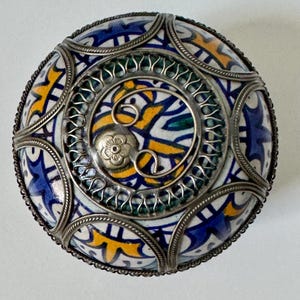 May include: A round, decorative ceramic box with a detailed, colorful pattern. The design features blue, yellow, and white geometric shapes, accented by a silver metal filigree border and a central spiral design. A small flower adorns the center.