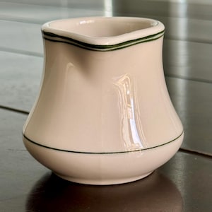 May include: Cream-colored ceramic creamer with a flared base and a spout. The creamer has a dark green trim around the top and bottom. The creamer is sitting on a dark brown surface.