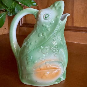 May include: A ceramic pitcher in the shape of a frog. The pitcher is primarily green with an orange belly and white accents. The handle is white with green shading. The frog's face is open, and the pitcher has a textured surface.