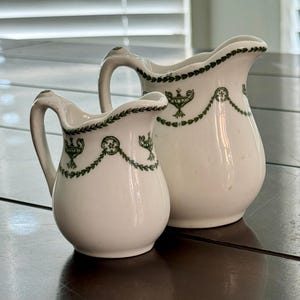 May include: Two white ceramic pitchers with green decorative trim. The pitchers have a classic design with a curved handle and a decorative green pattern around the rim and on the body. The pitchers are on a dark wooden surface.