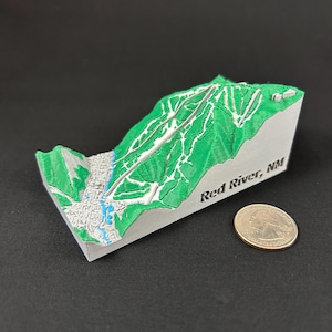 May include: A 3D-printed topographical map of Red River, NM, featuring green terrain with white ski runs and a blue river. The map is set on a gray base, with the text "Red River, NM" printed on the side. A quarter coin is next to the map.