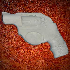 May include: A white plastic replica of a revolver handgun on a brown patterned background.