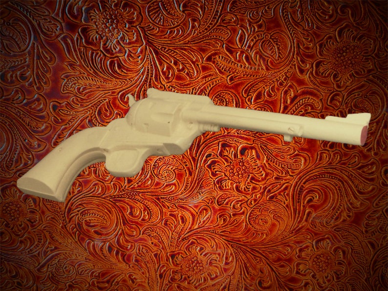 May include: A wooden toy gun, resembling a revolver, with a light brown finish. The gun is lying on a brown patterned surface.