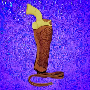 May include: A brown leather holster with floral embroidery, designed to hold a pistol. The holster is on a purple background with a floral pattern.