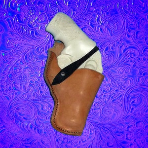 May include: A brown leather holster with a black strap for a white handgun. The holster is on a blue and purple patterned background.