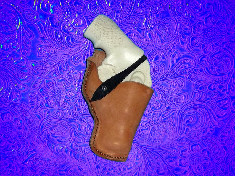 May include: A brown leather holster with a black strap for a white handgun. The holster is on a blue and purple patterned background.