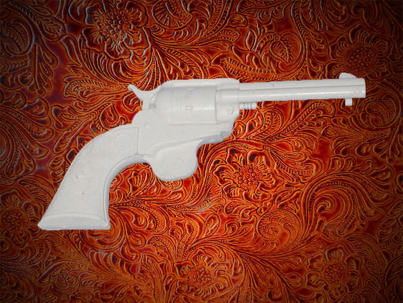 May include: A white plastic toy gun shaped like a revolver. The gun is lying on a brown leather surface with a floral pattern.