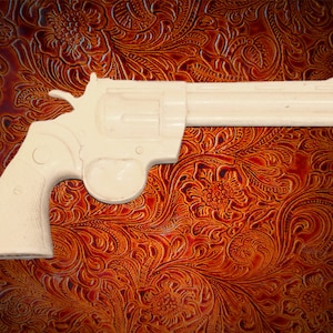 Può includere: Una pistola giocattolo di plastica bianca con un'impugnatura nera. La pistola è appoggiata su una superficie marrone con un motivo.