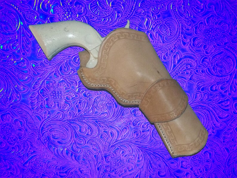 May include: A brown leather holster with intricate stitching and a white pistol inside. The holster is on a purple and blue patterned background.