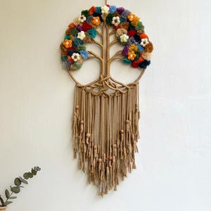 May include: A handcrafted wall hanging featuring a tree of life design. The tree has a circular crown adorned with colorful yarn flowers. The trunk and roots are made of woven rope, with hanging tassels and wooden beads. The wall decor is suspended from a natural fiber cord.