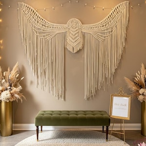 Op de afbeelding: Een beige macramé wandkleed in vleugelvorm hangt boven een groene fluwelen bank. De scène omvat gouden vazen met droge bloemstukken, een goudkleurig bord en een rond tapijt. De ruimte is goed verlicht.