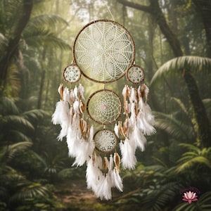 May include: A handcrafted dreamcatcher with a large central web and smaller circular webs. The dreamcatcher features a brown frame, cream-colored webs, and white and brown feathers. It is suspended against a blurred background of lush green foliage.