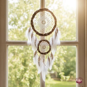 Puede incluir: Un atrapasueños artesanal con dos marcos circulares tejidos, adornado con plumas blancas y patrones de telaraña intrincados. El atrapasueños está hecho de materiales naturales y cuelga frente a una ventana con un fondo verde borroso.