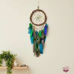 May include: A handmade dreamcatcher with a circular woven brown frame, a white web, and colourful beads. It features green, blue, and peacock feathers, creating a bohemian style. It hangs against a neutral wall, a decorative item.