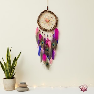 May include: A handcrafted dreamcatcher with a woven web design, adorned with pink, purple, and blue feathers, and peacock feathers. The dreamcatcher is suspended against a neutral background, creating a bohemian aesthetic.