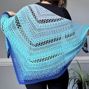 Pode incluir: Um xale de crochê azul e branco com um padrão de renda. O xale está drapeado sobre os ombros de uma pessoa e é feito com um fio degradê.