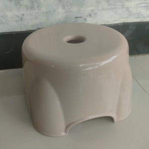 May include: A light beige plastic stool with a circular seat and a central hole. The stool has curved sides and a cutout at the base, offering a simple and functional design. The stool is suitable for indoor or outdoor use.