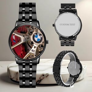 May include: Black wristwatch with a detailed face featuring a BMW car wheel design, including the BMW logo and red brake calipers. The watch has a black metal band and a custom text option on the back.