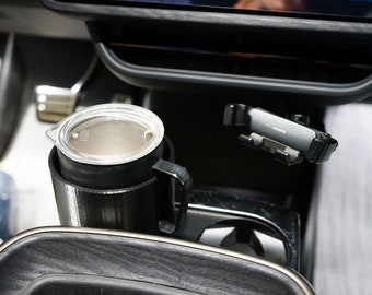 Op de afbeelding: Een zwarte en zilveren bekerhouder met een beker van roestvrij staal erin. De bekerhouder is bevestigd aan de middenconsole van een auto. Een zwarte telefoonhouder is bevestigd aan de zijkant van de console.