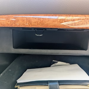 Peut inclure: Gros plan sur l'intérieur d'une voiture, montrant un petit compartiment de rangement avec une garniture en bois. Le compartiment est ouvert et vide, avec un loquet en plastique noir visible. Un document blanc est visible dans le coin inférieur droit de l'image.