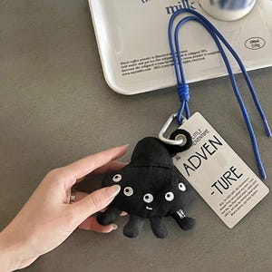 May include: Black plush alien-shaped keychain with white embroidered eyes and a small smile. Includes a blue lanyard and a tag that reads "LITTLE ADVENTURE". The keychain is held by a hand.