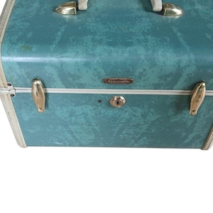 May include: Vintage teal Samsonite suitcase with a cream-colored handle and trim. The suitcase has gold-toned metal accents, including latches and a logo plate. The case features a keyhole lock and a textured surface.