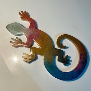 May include: A decorative lizard ornament in a vibrant gradient of colours. The lizard's body transitions from red to orange, yellow, green, and blue. It has a swirling pattern and a curled tail. The item is likely made of resin or a similar material.