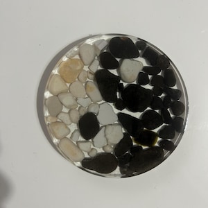 May include: A round coaster with a clear resin top. The coaster is divided into two sections, one with white and tan pebbles, and the other with black pebbles. The pebbles are of varying sizes and shapes.