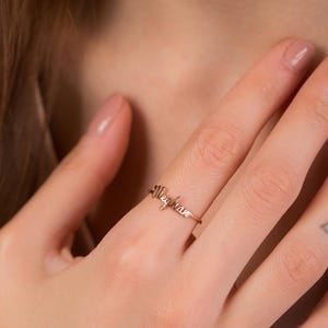 Può includere: Anello in oro rosa con il nome "Meghan" scritto in una delicata calligrafia. L'anello è indossato su un dito, mettendo in risalto il gioiello personalizzato. Il design dell'anello è semplice ed elegante, adatto all'uso quotidiano.