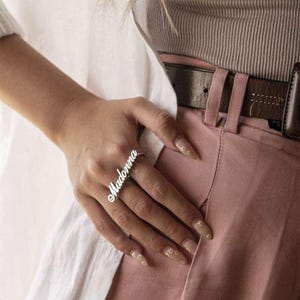 May include: A silver ring inscribed with the name "Madonna" in a cursive script. The ring is displayed on a finger, with a white shirt and pink trousers in the background. A personalized jewellery item.
