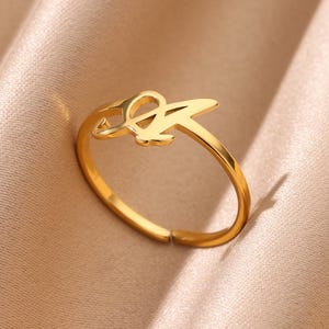 May include: A gold-colored ring featuring the letter "A" in a stylized script. The ring is adjustable and sits on a neutral-toned fabric. The ring is a piece of jewelry.