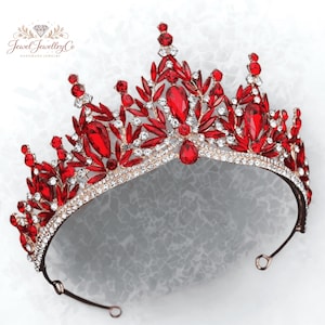 May include: A regal crown featuring a gold-toned base adorned with vibrant red gemstones and sparkling clear crystals. The crown's design includes leaf-like accents and teardrop-shaped gems, creating a luxurious and eye-catching accessory. The text "Jewel Jewellery Co" is visible.