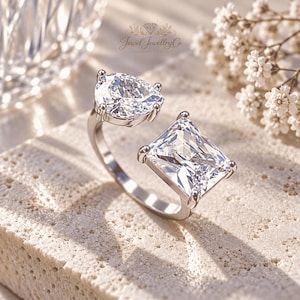 Pode incluir: Um anel aberto de tom prateado com duas pedras transparentes proeminentes, uma em forma de pera e outra retangular, montadas em garras. A banda é lisa e metálica. O texto Jewel JewelleryCo é visível na parte superior central da imagem.