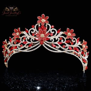 May include: A gold-toned tiara with red and clear gemstones. The tiara features floral and scrollwork designs, with red flower-shaped accents and clear crystal-like stones. The tiara is set against a black background.
