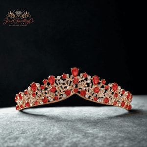 Op de afbeelding: Een tiara van roségoud met een symmetrisch, ingewikkeld filigraan metalen ontwerp, versierd met talloze heldere witte kristallen en levendige rode druppelvormige stenen. Het hoofddeksel heeft een puntig midden en een gebogen structuur.