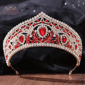 May include: A gold-toned crown with red and clear gemstones. The crown features a central teardrop-shaped red gem, surrounded by smaller red and clear stones. The design includes leaf-like accents and a curved base, suitable for special occasions.
